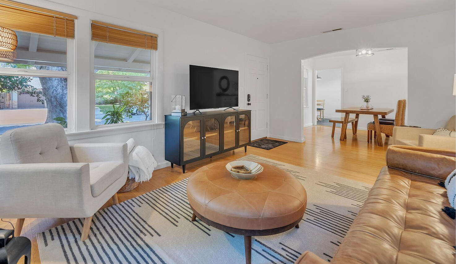 This mid-century modern family room exudes timeless elegance with its open layout, featuring a sleek black media console housing with SmartTV. The low-profile genuine leather sofa, paired with classic accent chairs, offers comfortable seating. A stylish ge